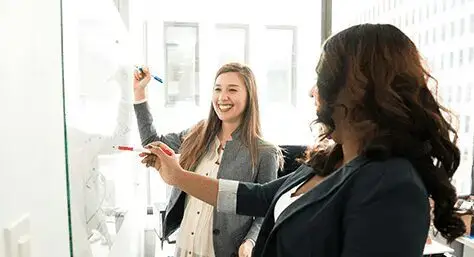 Two women are smiling while one of them is writing on a board.